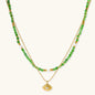 Layered necklace with green stone beads, gold spacers, white accents, and protective eye pendant on cream background.