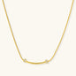 Delicate gold-plated necklace with curved bar pendant and crystal accents on snake chain against cream background.
